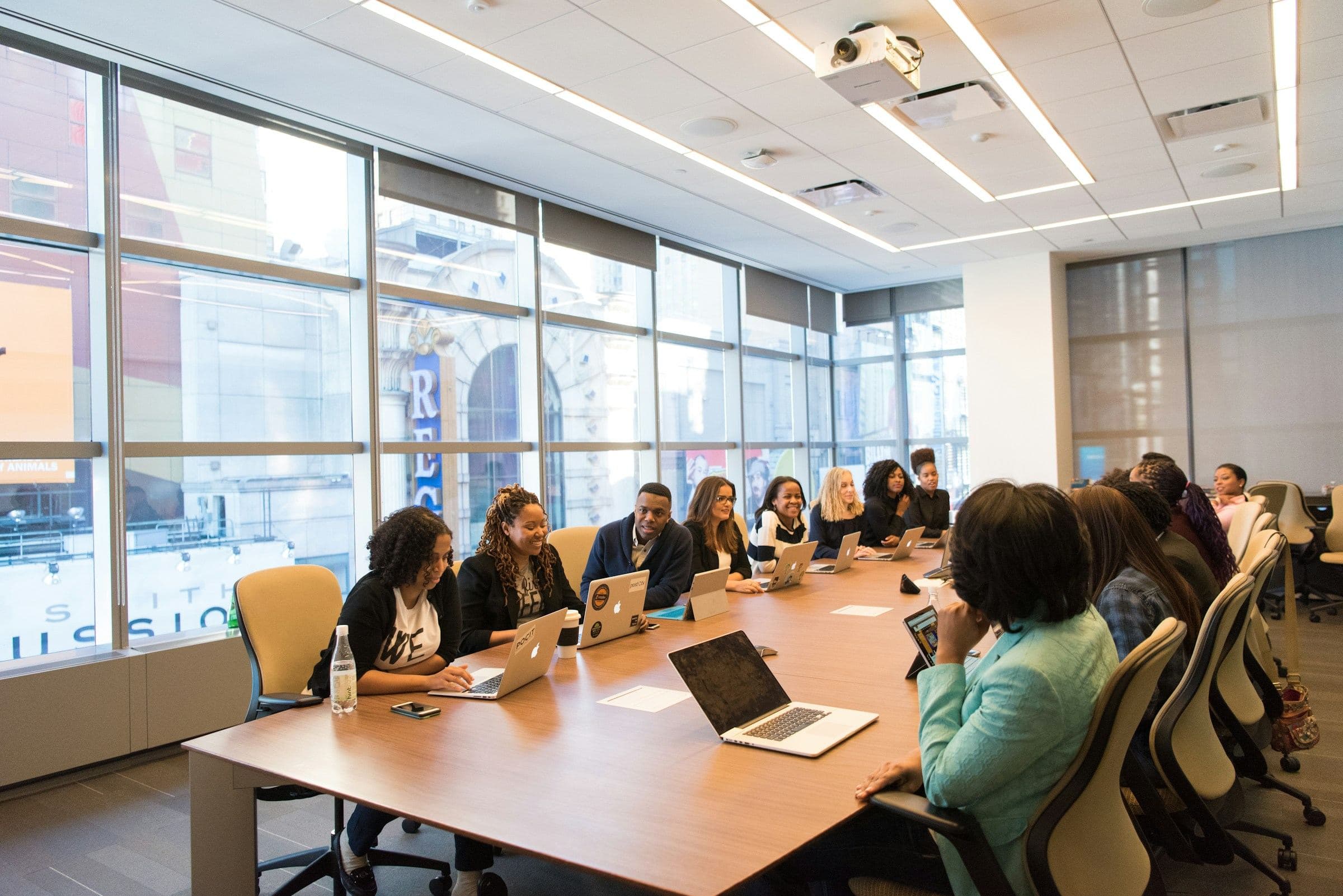 A large team seated along a boardroom table with laptops, smiling during a meeting